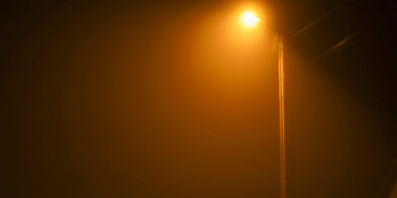 A bright orange streetlight on a tall pole pierces through thick fog or haze on a dark night. The light creates a halo, giving the scene a mystical and enigmatic appearance.