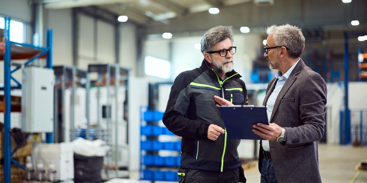 Two colleagues engage in a professional discussion inside a modern industrial warehouse.