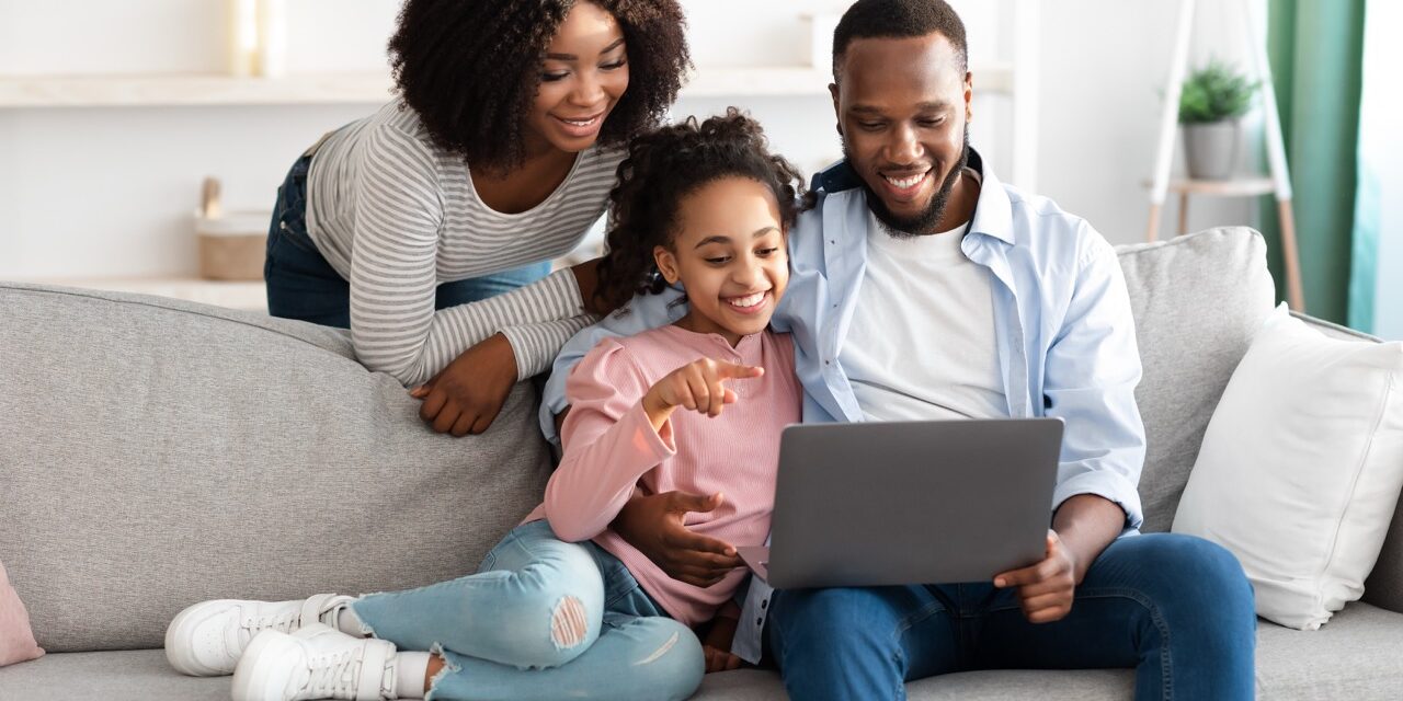 People And Technology Concept. Portrait of cheerful African American family of three using laptop, dad hugging daughter, teenage girl pointing at screen, showing photos or product in online webstore